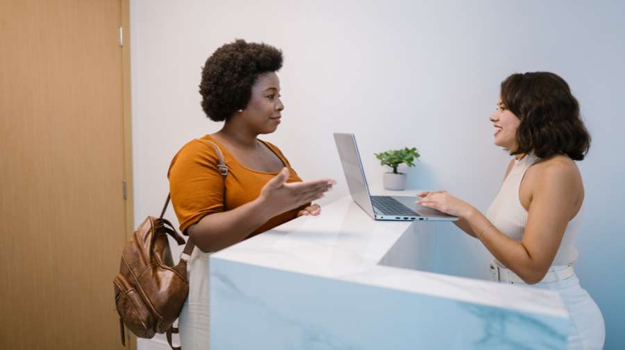 two women sitting across from each other at a desk with a laptop