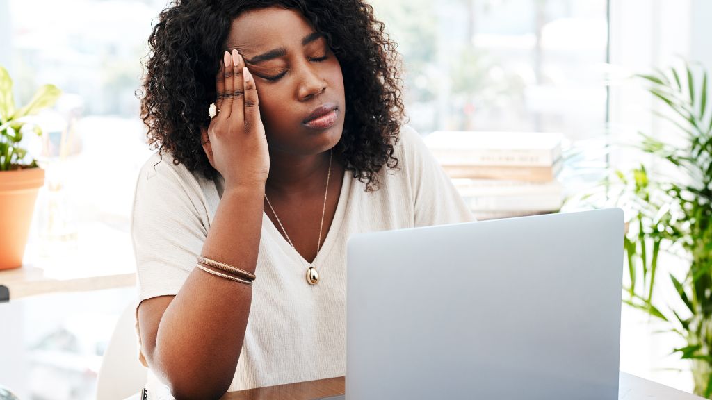 woman sitting in front of laptop with closed eyes and her hand on her temple, looking tired and frustrated