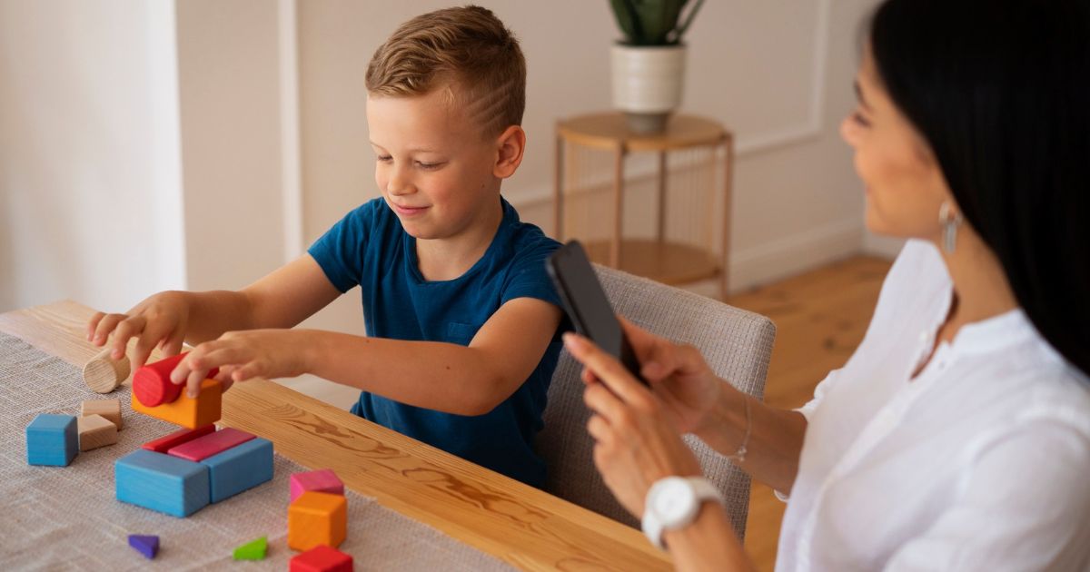 woman using a phone to take notes as she watches a boy complete a wooden block structure on the table in front of him