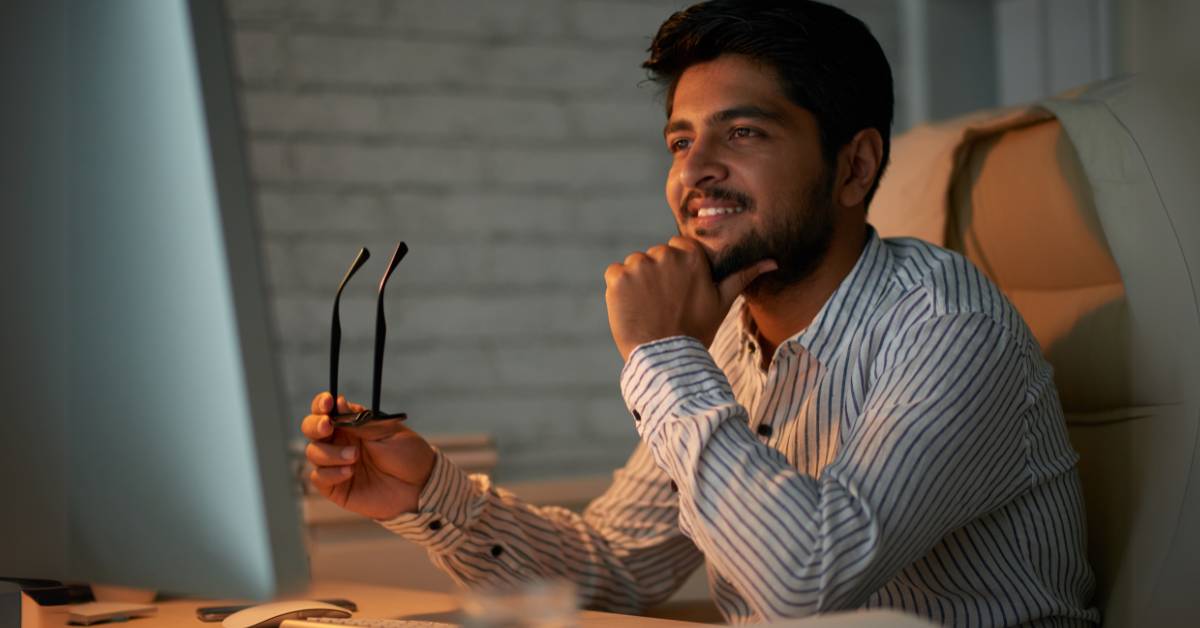 man sitting at a desk, holding glasses and smiling